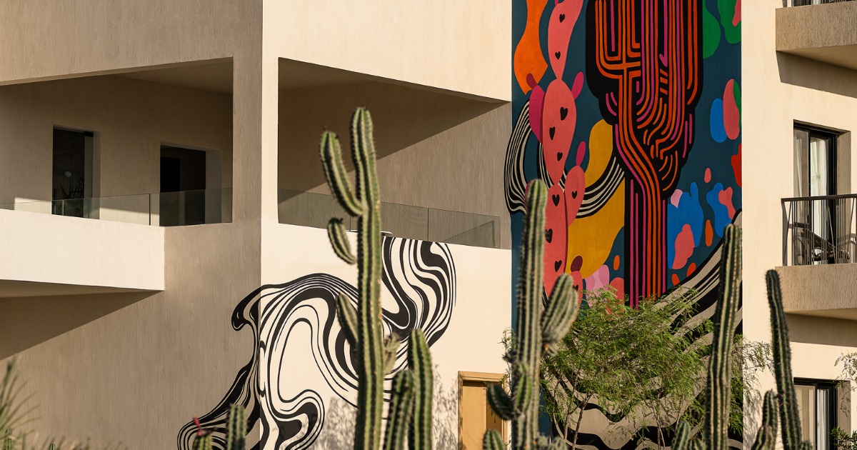 Close-up of colourful large-scale street art murals on the hotel exterior facade, framed by tall cacti in the foreground