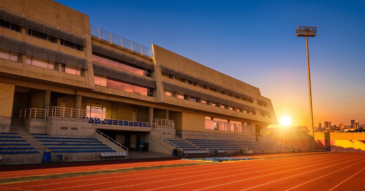 Stadium grandstand and 8-lane running track at sunset, with a floodlight tower illuminating the straight