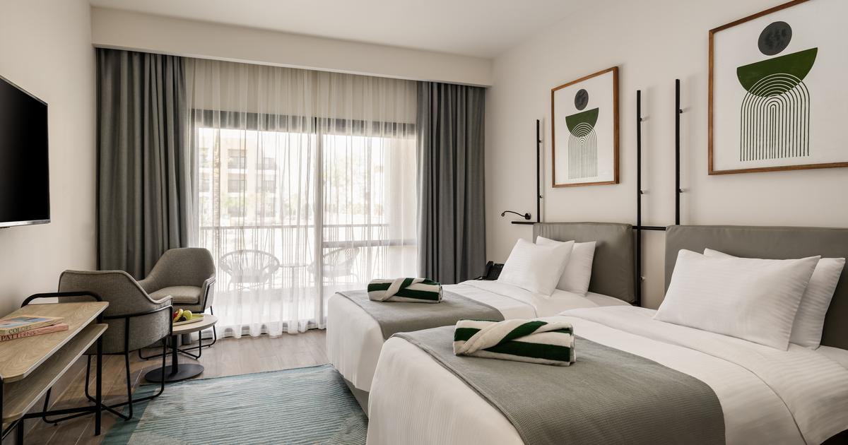 Twin-bed hotel room with neutral tones, balcony access, wall-mounted artwork, and a flat-screen TV