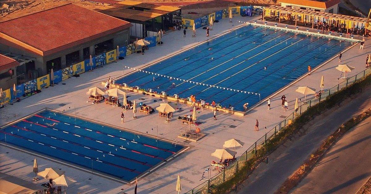 Aerial golden-hour view of a multi-pool swimming facility with lane ropes, umbrella shades, and coaches on deck