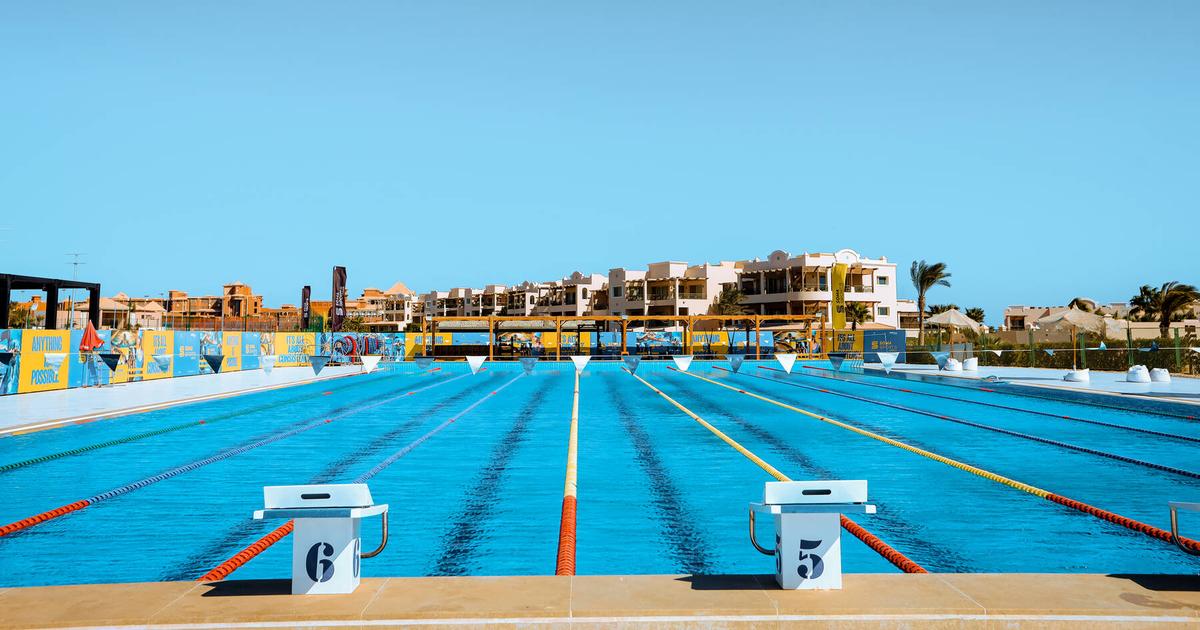 Competition swimming pool viewed from starting blocks 5 and 6, with resort buildings and sponsor banners in the background