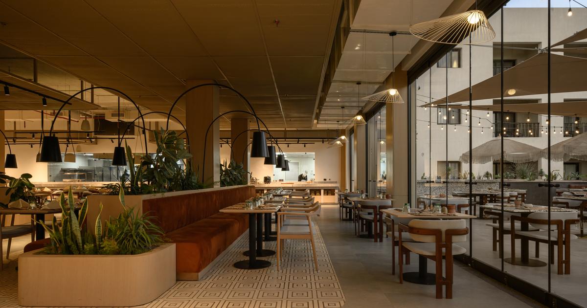 Modern hotel restaurant interior with warm lighting, banquette seating, tropical plants, and floor-to-ceiling windows overlooking a courtyard