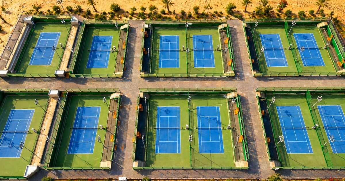 Дневная аэрофотосъёмка шести синих теннисных кортов ITF в Сома-Бей, расположенных в два ряда, в окружении пустынного песка и пальм